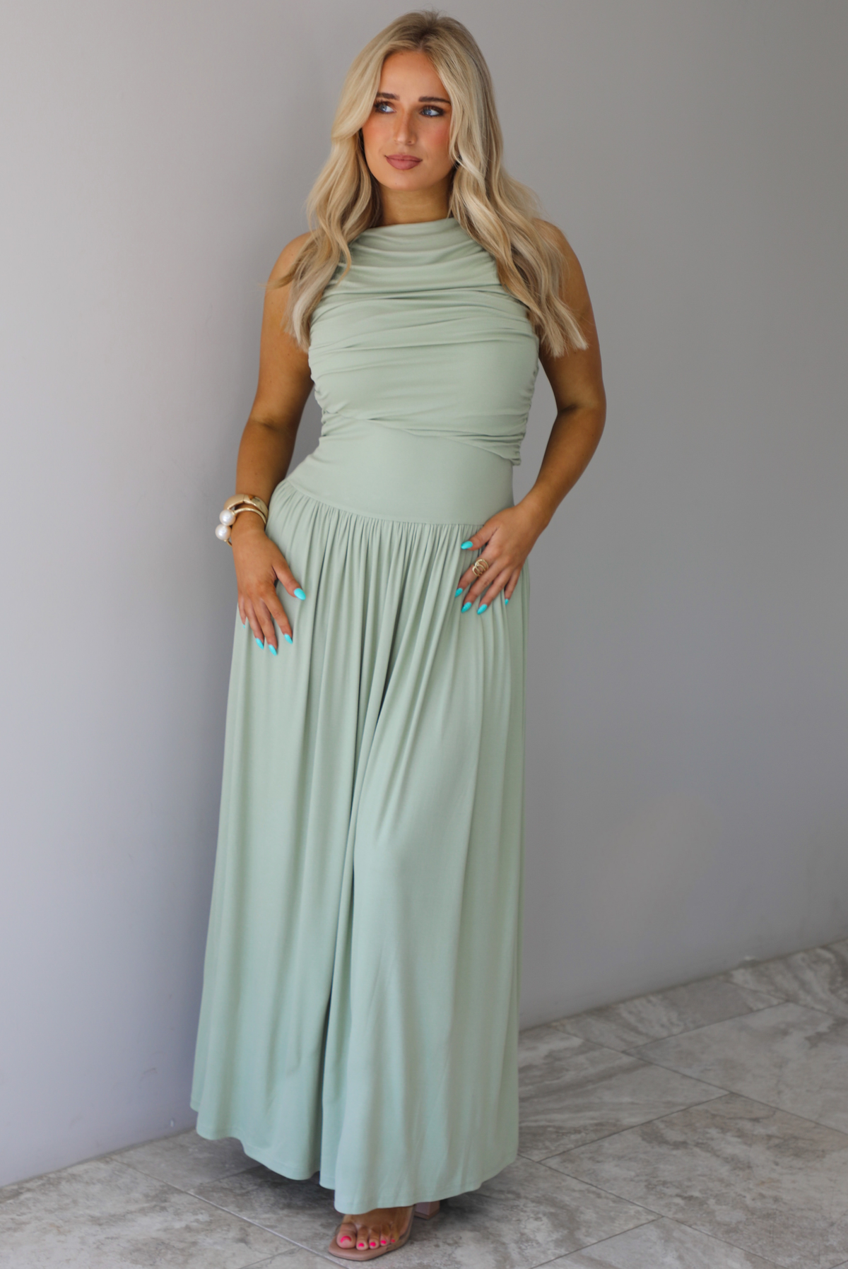 Woman wearing a sage green maxi length dress, which has a rouched detail at the bust, a fitted waistline, a flowy skirt. The photo is taken indoors against a white wall with white marble flooring. 