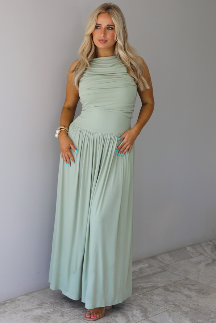 Woman wearing a sage green maxi length dress, which has a rouched detail at the bust, a fitted waistline, a flowy skirt. The photo is taken indoors against a white wall with white marble flooring. 
