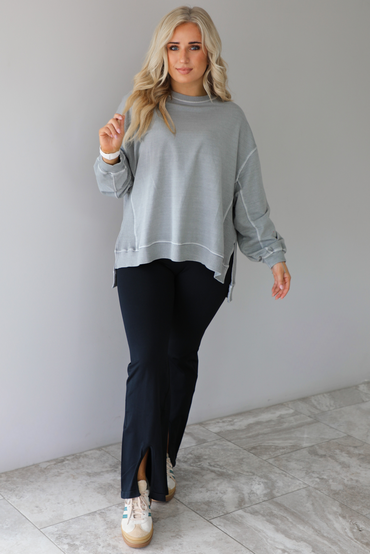 Woman wearing a grey oversized sweater with black leggings that have a slightly flared hem with split hem details in the front. The photo is taken in front of a white wall with white marble flooring. 