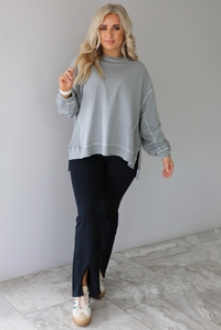 Woman wearing a grey oversized sweater with black leggings that have a slightly flared hem with split hem details in the front. The photo is taken in front of a white wall with white marble flooring. 