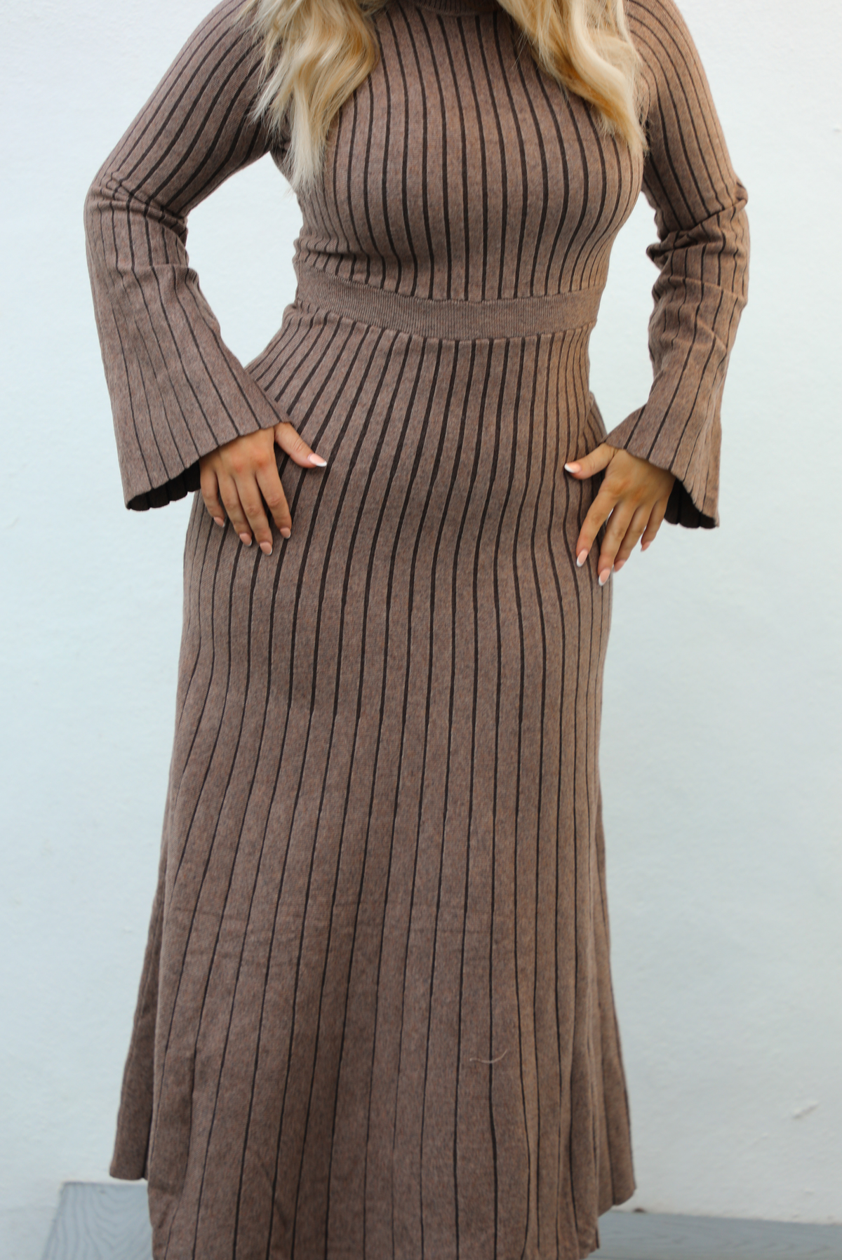 Woman wearing a taupe colored dress that has a vertical ribbed pattern throughout, a high neckline, fitted bodice, cinched waistline, long sleeves that flare slightly, and a maxi length skirt that flows at the bottom. The photo is taken outdoors against a white wall.  The photo shows a close up of the dress front the neckline, down.