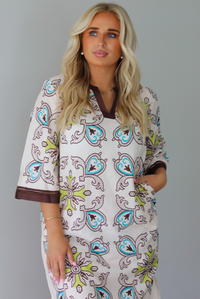 A woman modeling a long kaftan dress with a colorful paisley pattern on a cream background, featuring brown trim and sandals, standing indoors against a white wall.