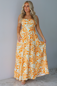A woman modeling a spaghetti strapped, floor-length maxi dress with a white base and a vibrant marigold orange floral pattern, standing indoors against a white wall with white marble flooring. 