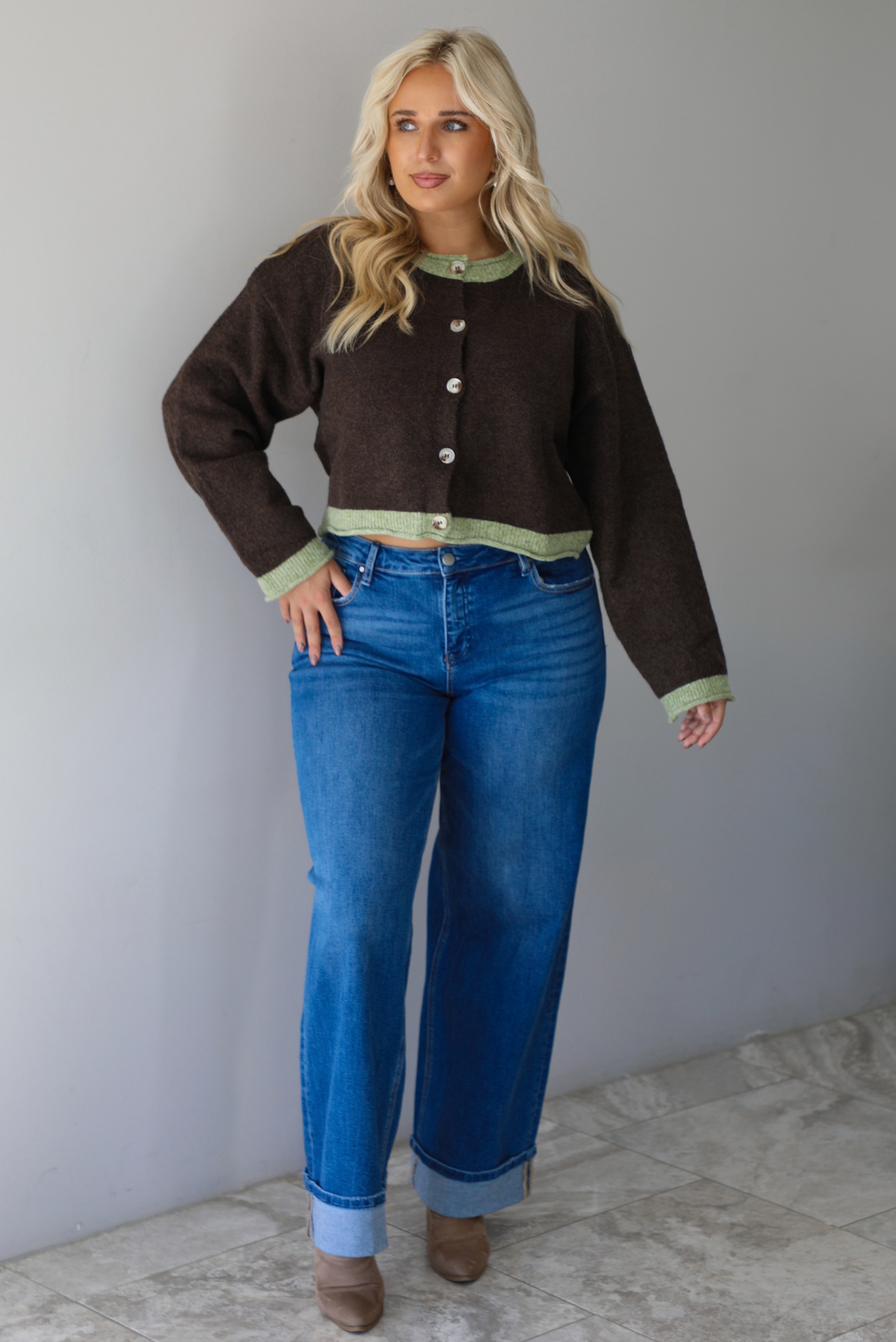 A woman wearing a dark brown with green trim cardigan fully buttoned up paired with dark wash, cuffed hem denim jeans, standing indoors against a white wall with white marble flooring. 