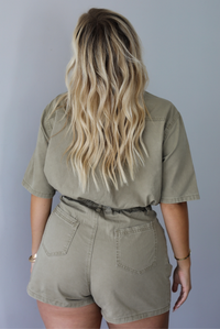 A woman shown from behind modeling a short-sleeve khaki jumpsuit with a button-front collared romper with a cinched waist, standing indoors against a white wall.