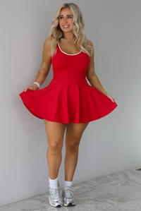 Woman wearing a red athleisure dress that has white trim at the top of the bodice, a fitted bodice, and flowy skirt. The dress is paired with high white ankle socks and silver sneakers. The photo is taken indoors against a white wall with white marble flooring. 