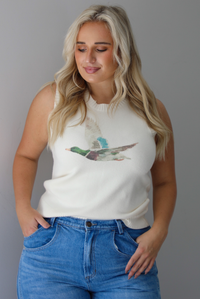 A woman wearing a cream colored sweater vest with a mallard duck on the front, paired with medium wash denim with wide leg hem, standing indoors against a white wall.