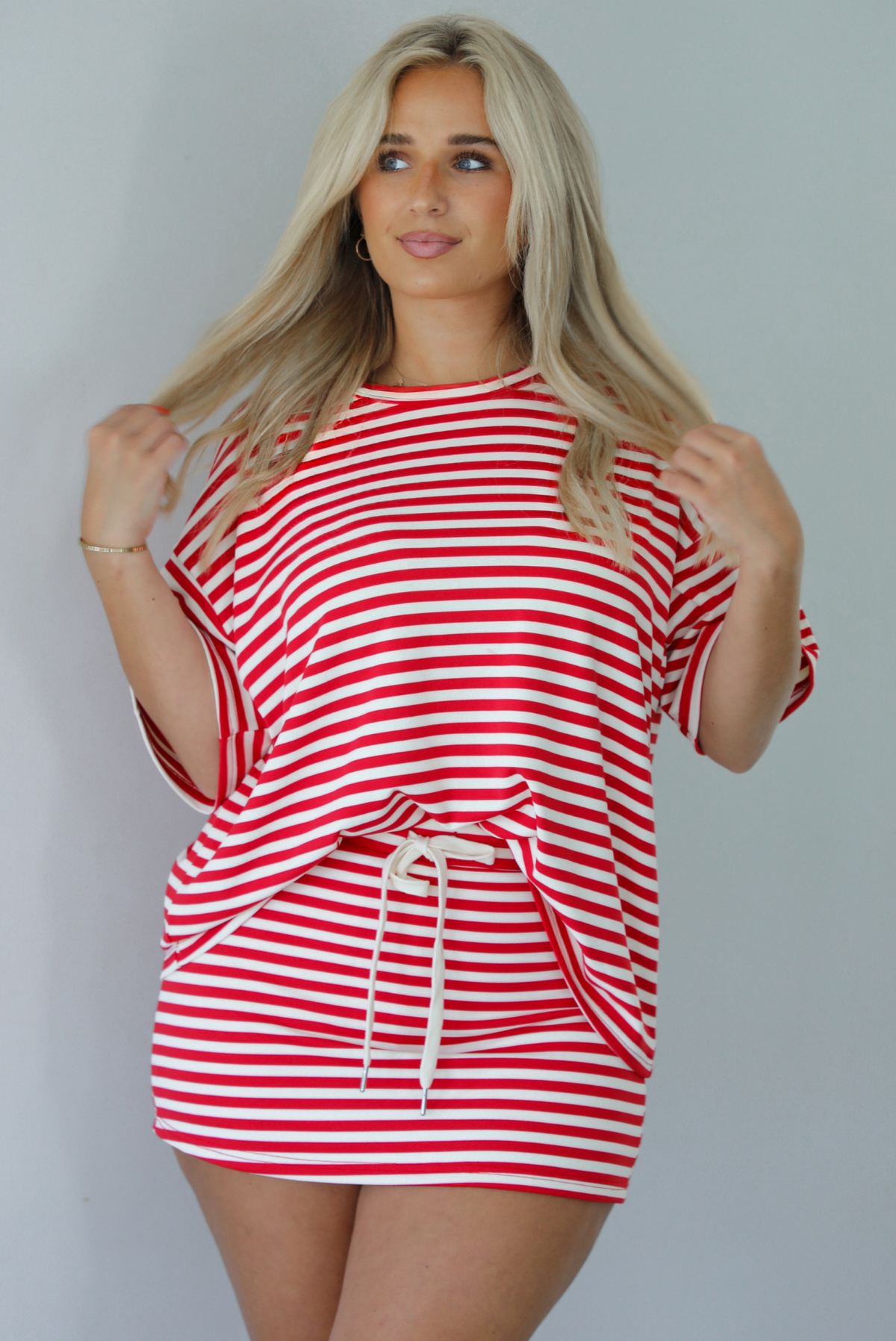 Woman wearing a red and white striped set featuring an oversized top and mini skirt indoors against a white wall. The photo shows a closeup of the set and has the top tucked into the skirt, showing that it has a drawstring waistline. 