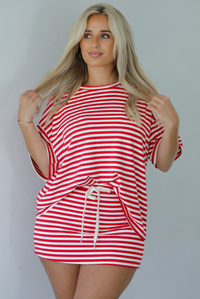 Woman wearing a red and white striped set featuring an oversized top and mini skirt indoors against a white wall. The photo shows a closeup of the set and has the top tucked into the skirt, showing that it has a drawstring waistline. 