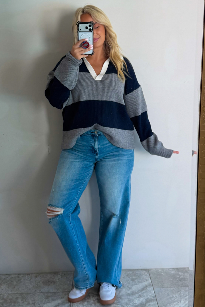 A woman taking a mirror selfie wearing a navy and charcoal boyfriend style pullover top with a white collared detail, with distressed denim, standing indoors against a white wall with white marble flooring. 