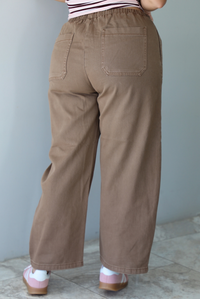 A closeup of the back of a woman wearing mocha barrel pants that have an elastic and drawstring waistline, paired with a pink, burgundy, and white striped design and pink sneakers, standing indoors against a white wall with white marble flooring. 