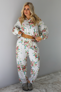 Woman wearing an oatmeal colored and multi-colored floral print two piece set featuring a collared long sleeved crop top and high waisted pants. The photo is taken indoors against a white wall with white marble flooring. 