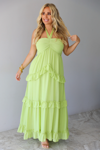 A woman modeling a lime green halter-neck maxi dress with tiered ruffle details, standing indoors against a white wall with white marble flooring. 