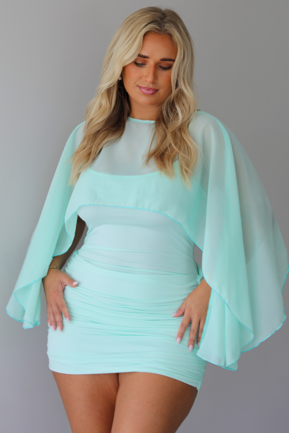 A woman in a light mint green mini dress with a sheer draped shawl, standing indoor against a white wall.