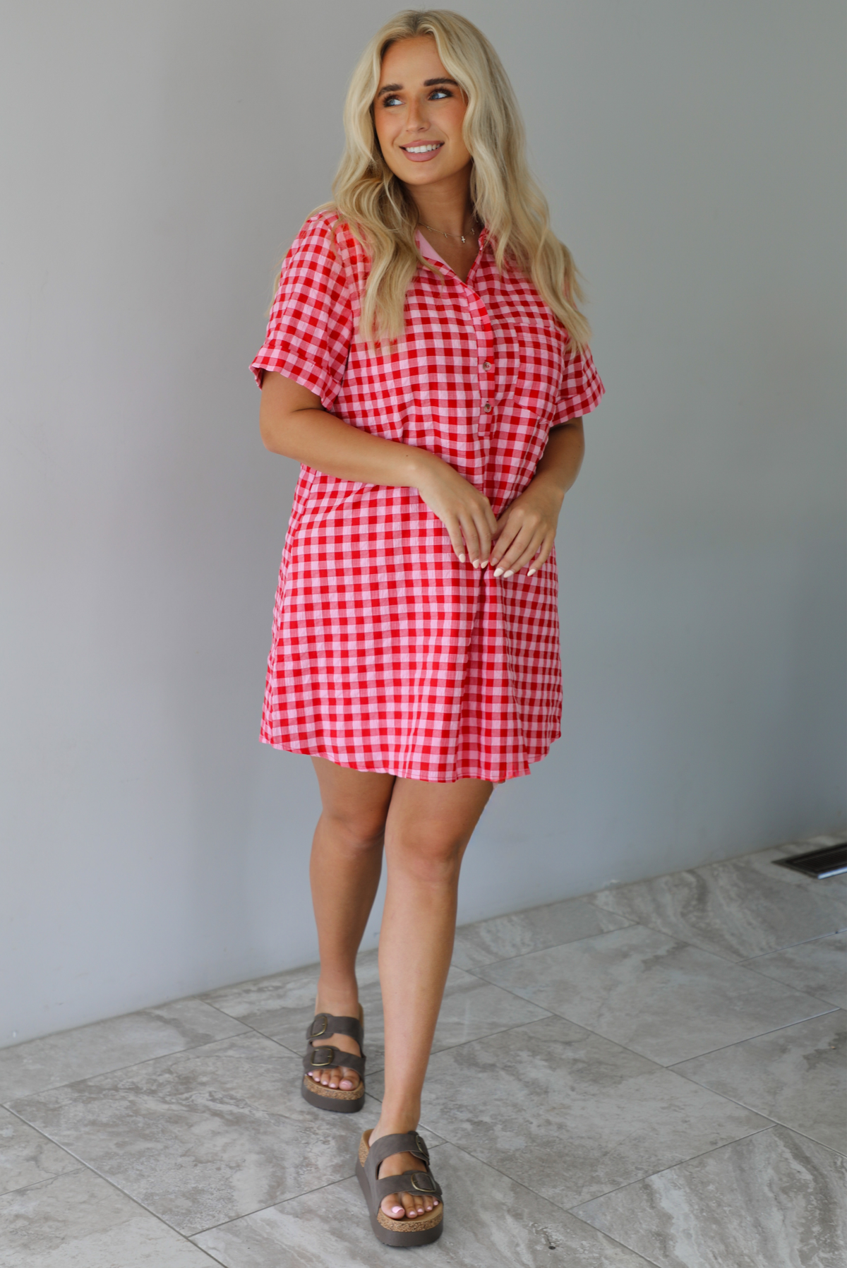 Woman wearing a pink and red gingham collared neckline t-shirt dress indoors against a white wall with white marble flooring. 