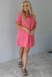 Woman wearing a pink and red gingham collared neckline t-shirt dress indoors against a white wall with white marble flooring. 