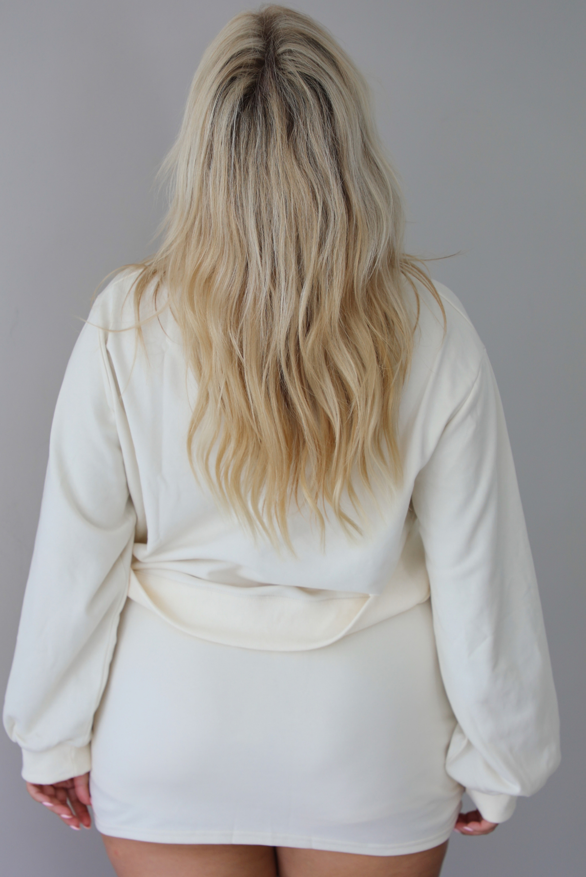 A woman shown from behind wearing a cream matching two piece set featuring a cropped sweatshirt and mini skirt of the same material, standing indoors against a white wall. The front of the sweater is untucked to show the full length. 