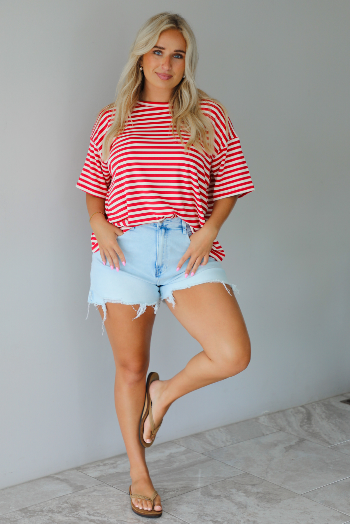 A woman modeling a red and white horizontally striped short-sleeve t-shirt with light blue denim shorts and brown sandals, standing indoors against a white wall with white marble flooring. 