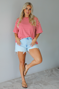 A woman modeling a red and white horizontally striped short-sleeve t-shirt with light blue denim shorts and brown sandals, standing indoors against a white wall with white marble flooring. 