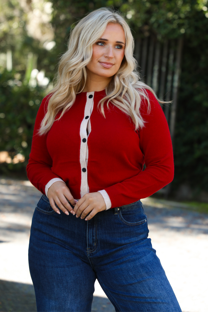 Woman model wearing a red cardigan with sand trim details and black button closures in the front with dark wash denim. The photo shows a close up of the cardigan. The photo is taken outdoors on gravel with greenery in the back. 