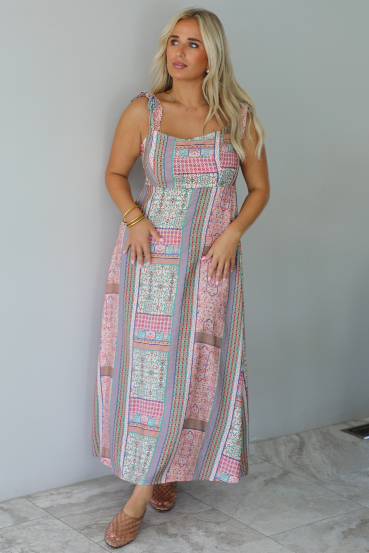 A woman wearing a thick strapped maxi dress with a patchwork print of pink gingham, beige paisley, and blue patterns, standing indoors against a white wall with white marble flooring. 