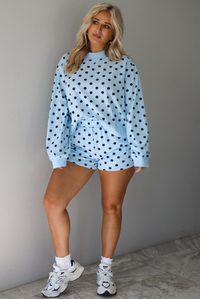 A woman wearing a blue two piece set with a navy polka dot pattern throughout, featuring a sweatshirt and high waisted sweat-shorts, paired with chunky sneakers and high ankle socks, shown indoors against a white wall with white marble flooring. 