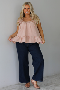 A woman wearing a light maroon and cream gingham flowy top with dark denim jeans, standing indoors against a white wall with white marble flooring.