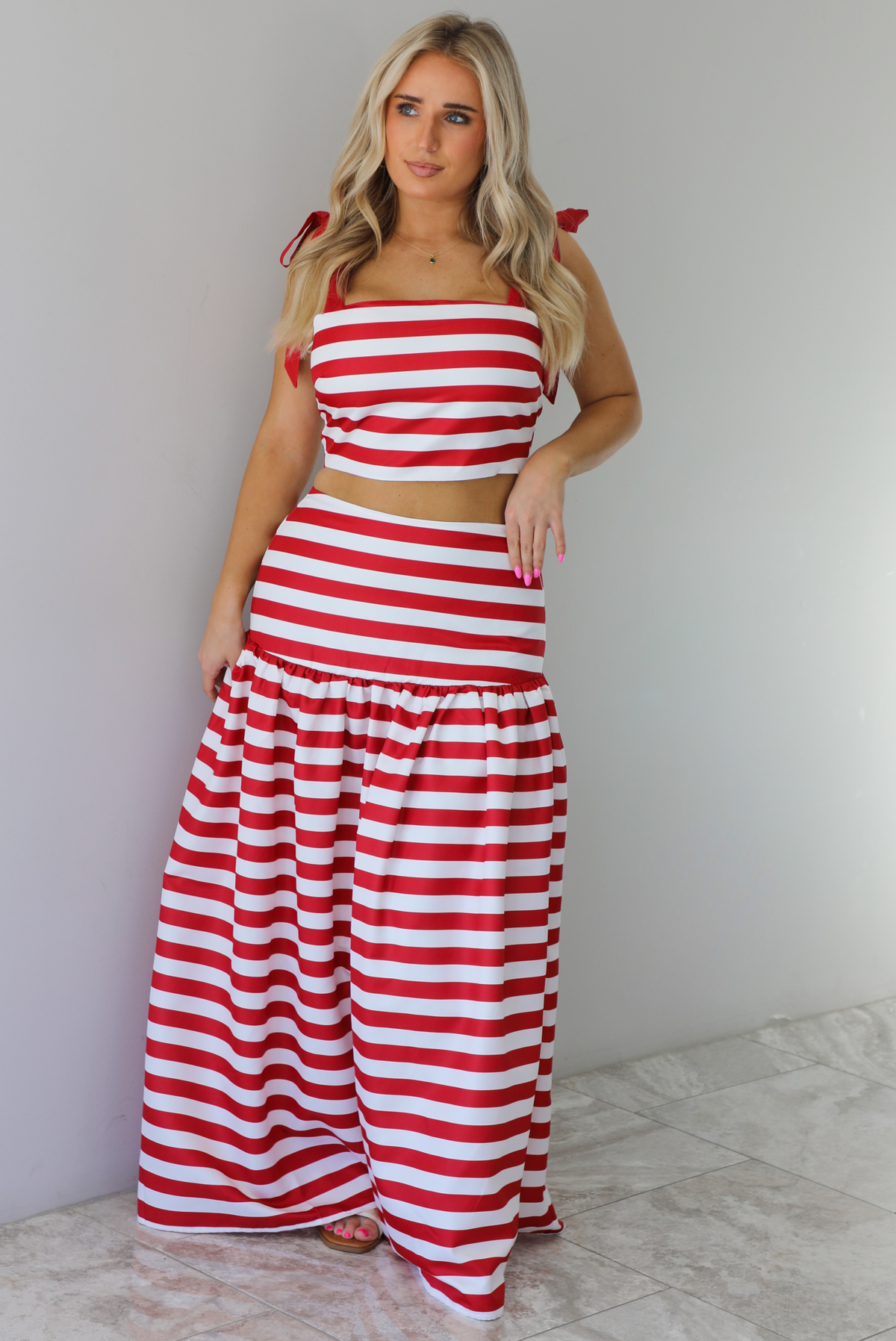 Woman wearing a red and white striped two piece set featuring a cropped top and maxi length, high waisted skirt. The photo is taken indoors against a white wall with white marble flooring. 