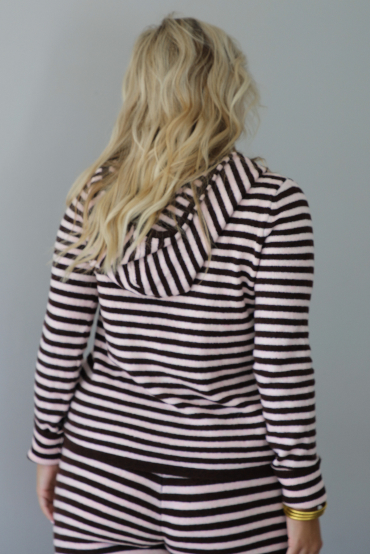A woman wearing a light pink and brown striped two piece set featuring a double zipped hoodie top and mid-rise, wide leg pants, standing indoors against a white wall. The photo shows a closeup of the back of the top and its hoodie detail. 