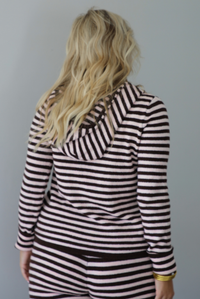 A woman wearing a light pink and brown striped two piece set featuring a double zipped hoodie top and mid-rise, wide leg pants, standing indoors against a white wall. The photo shows a closeup of the back of the top and its hoodie detail. 