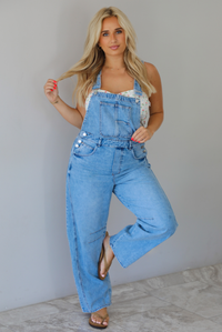 Woman wearing a pair of denim overalls with a white floral strapless top underneath indoors against a white wall with white marble flooring. 