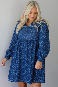 A woman wearing a dark denim mini dress with a white floral print pattern throughout, collared neckline, a quilted detail at the bodice, long sleeves, and a flowy skirt, standing indoors against a white wall.