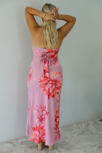 A woman viewed from behind wearing a strapless mauve maxi dress with a large red and orange floral print and a tie detail at the back waist, standing indoors against a white wall with white marble flooring. 