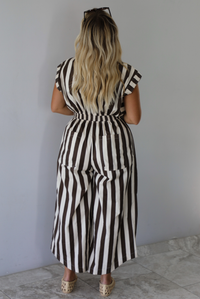 A woman shown from behind wearing a brown and white striped patterned jumpsuit with a collared neckline, functional button closures in the front, a cinched waistline, and barrel-style bottoms, standing indoors against a white wall with white marble flooring. 