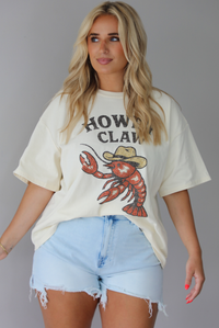 Woman wearing a graphic t-shirt with a lobster wearing a cowboy hat and the words 'HOWDY CLAW' written along the front and denim shorts against a white wall. The photo shows the tee front tucked into the denim shorts. 