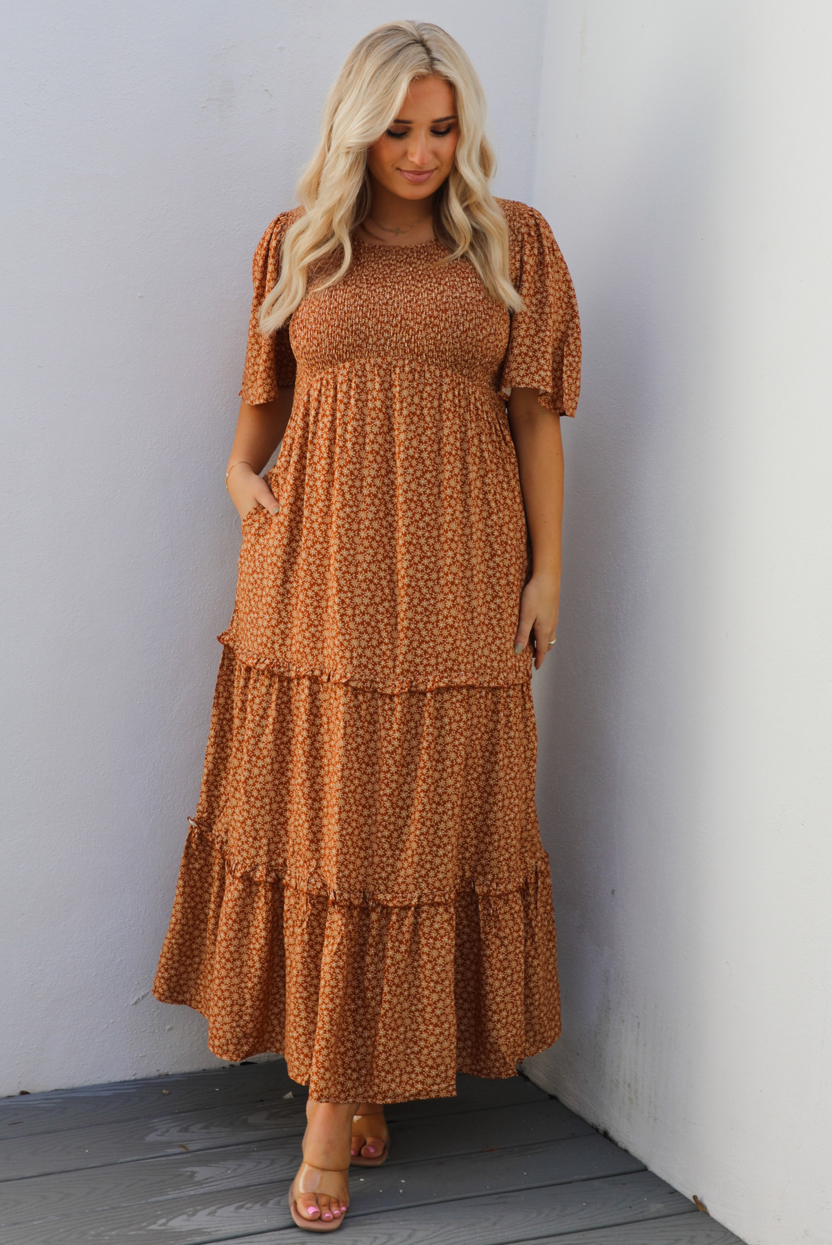 A woman model outdoors wearing a rust dress with a cream floral print pattern throughout. It has flowy short sleeves, a smocked bodice, and a flowy maxi length skirt with functional pockets on either side. 