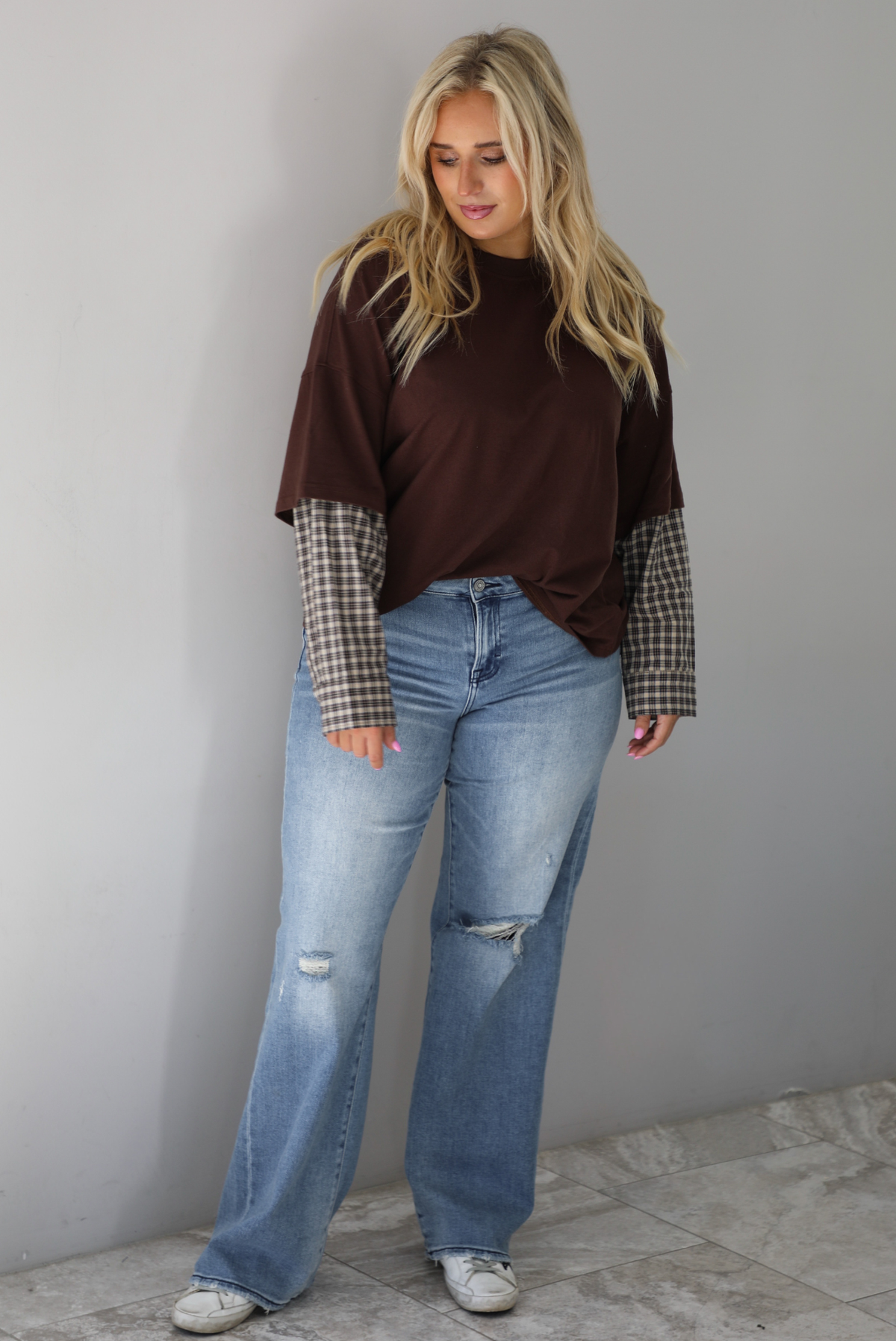 A woman wearing a brown t-shirt with a layered look of plaid sleeve detail, paired with light wash denim jeans, standing indoors against a white wall with white marble flooring. 