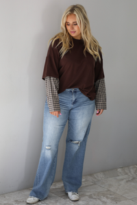 A woman wearing a brown t-shirt with a layered look of plaid sleeve detail, paired with light wash denim jeans, standing indoors against a white wall with white marble flooring. 