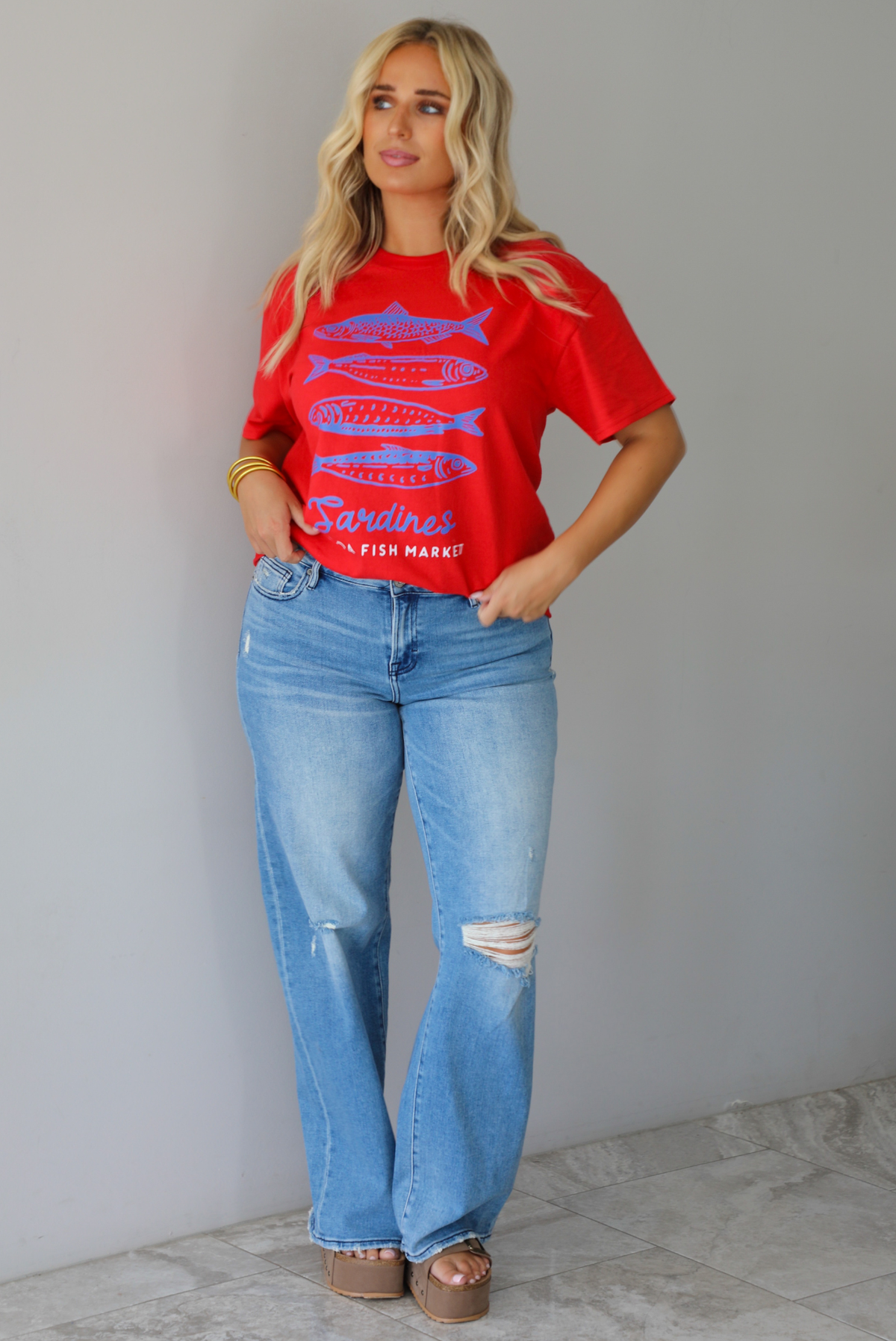 A woman wearing a red graphic t-shirt with blue sardine illustrations and the text 'Sardines LISBON FISH MARKET', paired with blue jeans, standing indoors against a white wall with white marble flooring.