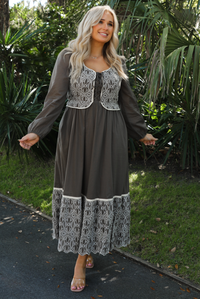 Woman model wearing an olive and off-white stitched detail dress outdoors. Dress has a corset-appearance bodice, long sleeves, functional pockets on either side, an elastic waistline, and a long, maxi length.
