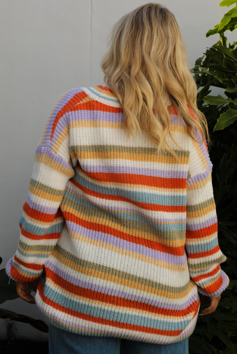 Woman model wearing an orange, lavender, yellow, muted green, blue, and white striped oversized cardigan with functional button closures in the front and an oversized fit with a light wash flared hem denim. The photo is taken outdoors against a white wall and greenery in the back. The photo showcases a close up of the back of a cardigan.