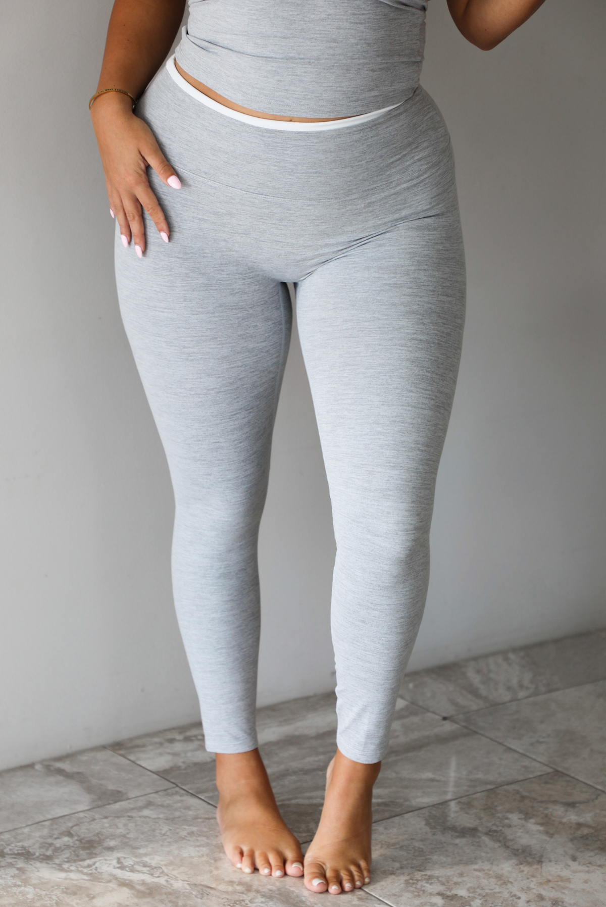 A woman modeling a light gray two-piece loungewear set consisting of a cropped tank top and matching leggings, focused on a closeup of the pants, standing indoors against a white wall with white marble flooring. 