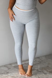 A woman modeling a light gray two-piece loungewear set consisting of a cropped tank top and matching leggings, focused on a closeup of the pants, standing indoors against a white wall with white marble flooring. 