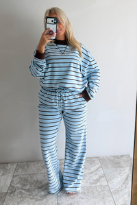 A woman wearing a blue matching two piece set with dark brown and white striped details, featuring an oversized long sleeved top and high waisted loose fitting, flared leg pants. She is taking a mirror selfie in front of a white wall with white marble flooring. 