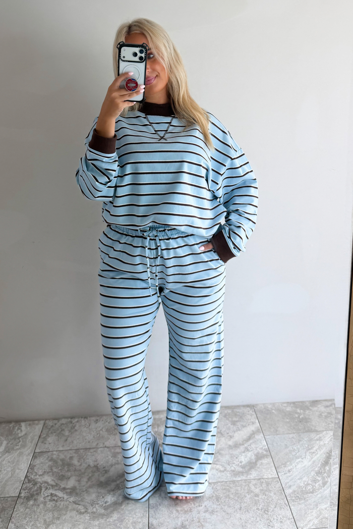 A woman wearing a blue matching two piece set with dark brown and white striped details, featuring an oversized long sleeved top and high waisted loose fitting, flared leg pants. She is taking a mirror selfie in front of a white wall with white marble flooring. 