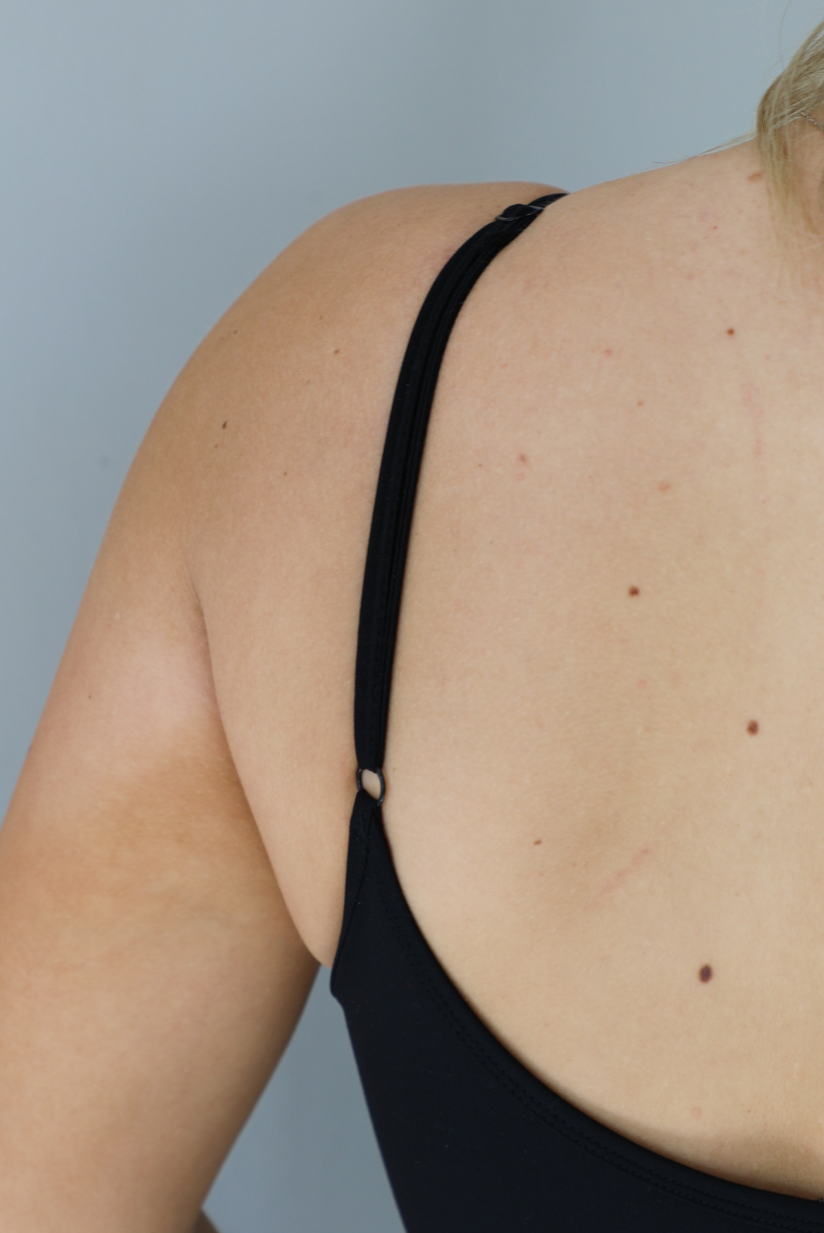 Woman wearing a black fitted jumpsuit with spaghetti straps. The jumpsuit is styled with pink leg warmers. The photo shows a closeup of the adjustable strap on the jumpsuit. The photo is taken in front of a white wall indoors. 