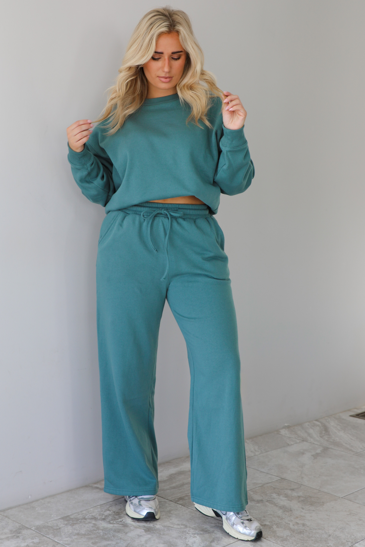 Woman wearing a sage two piece sweat-set. The set features a slightly oversized sweater and high waisted sweat pants that have an elastic and drawstring waistline with pockets. The photo is taken indoors against a white wall with white marble flooring. 