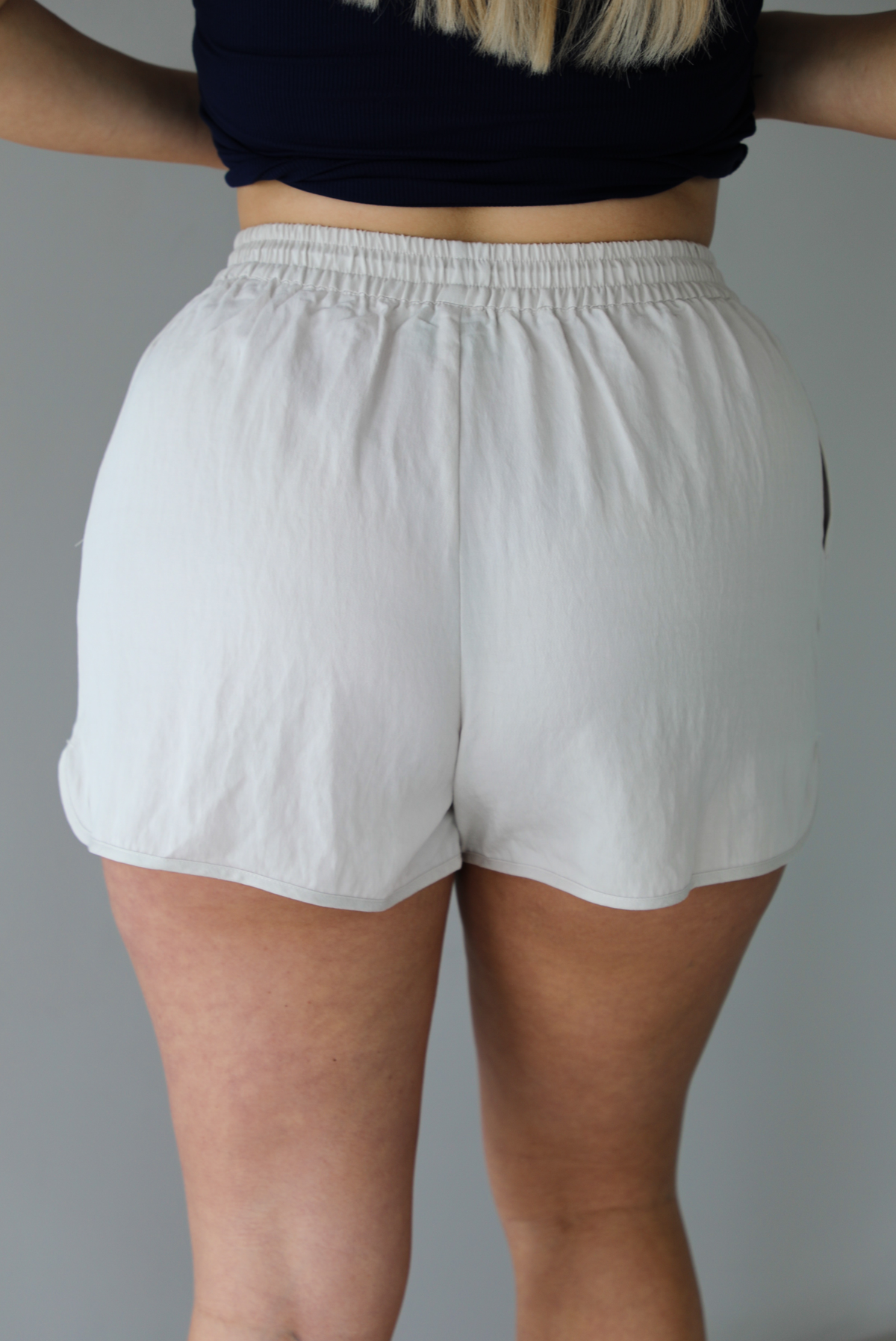 Woman wearing a stone colored two piece set featuring a zip up jacket and shorts with a navy tank tucked into the shorts indoors against a white wall. The photo shows a closeup of the back of the shorts. 