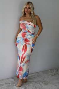 A woman modeling a strapless off-the-shoulder maxi dress with a vibrant red, blue, and cream floral print against a white wall with white marble flooring. 
