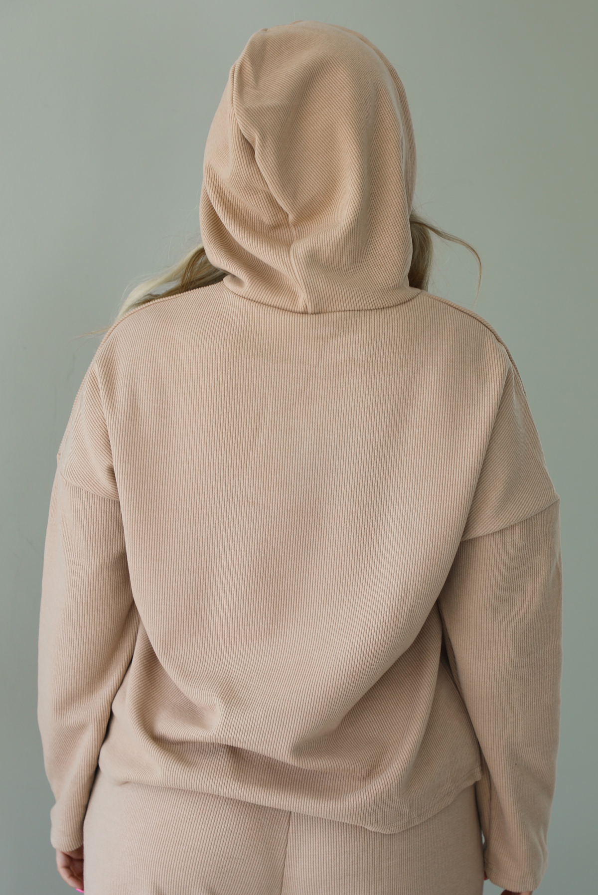 Woman wearing a tan two piece set with a hoodie top and sweatpants. The photo shows the back of the hoodie. The photo is taken indoors against a white wall.